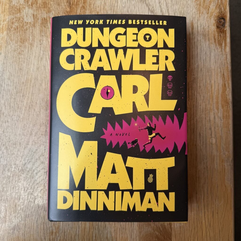 A hardcover copy of Dungeon Crawler Carl by Matt Dinniman sitting on a light brown wooden table with the front cover facing the camera.