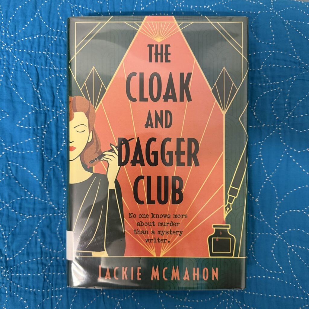 A hardcover copy of The Cloak and Dagger Club by Jackie McMahon sitting on a blue cloth surface. The front cover is facing the camera.