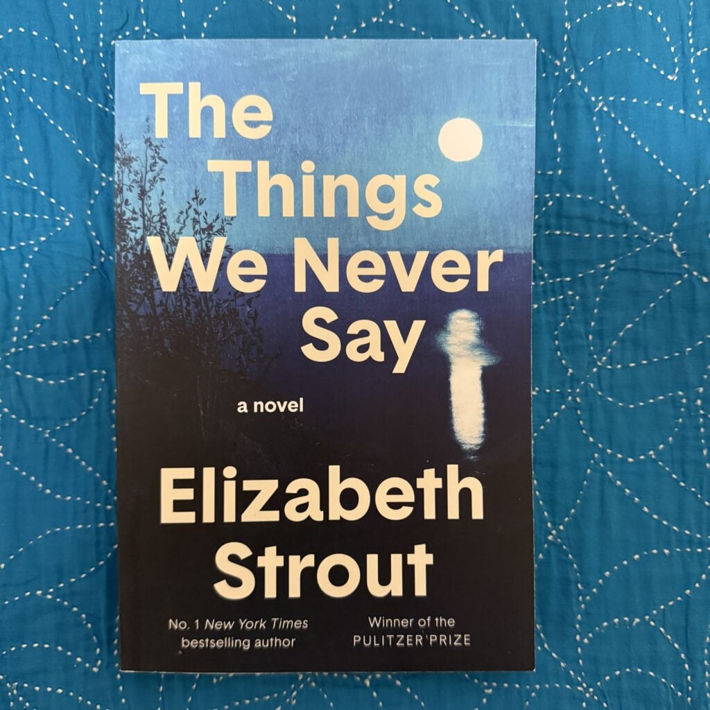 A paperback copy of The Things We Never Say by Elizabeth Strout on a blue cloth surface with the front cover facing the camera.