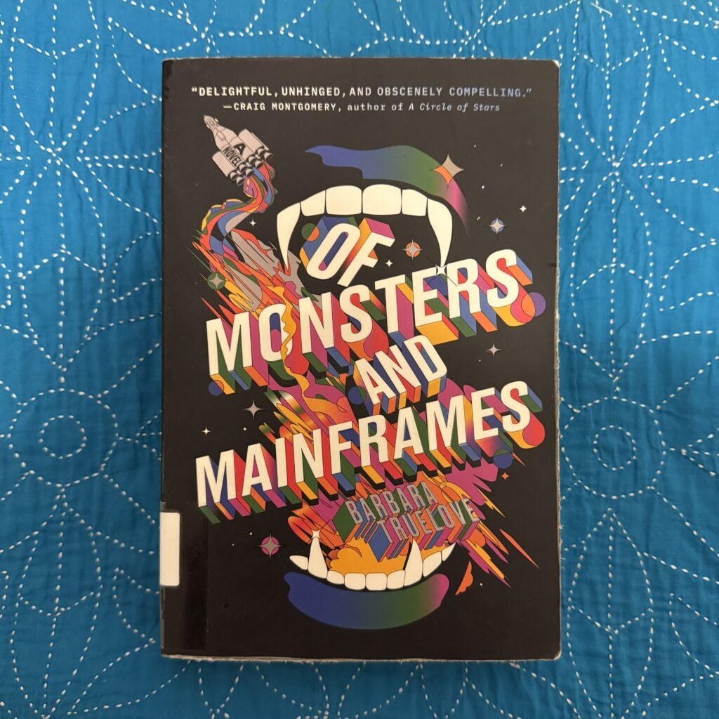 A picture of a paperback copy of Of Monsters and Mainframes by Barbara Truelove sitting on a blue cloth surfaces with the front cover facing the camera.