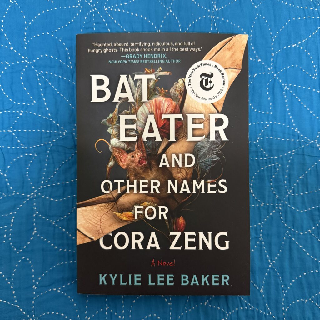 A picture showing my paperback copy of Bat Eater and Other Names for Cora Zeng by Kylie Lee Baker sitting on a blue cloth surface. The front cover is facing the camera.